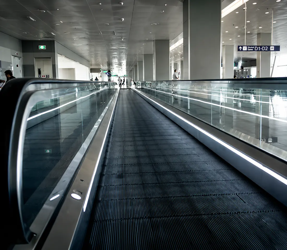 An empty travelator stretches toward an illuminated gate directory sign in an airport concourse.