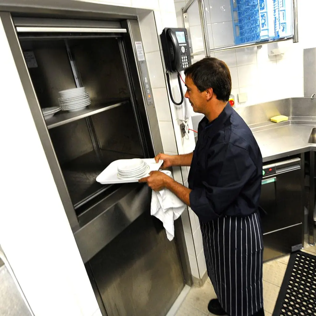 Stainless steel service lift standing open in a restaurant kitchen, its shelf loaded with a chafing dish and stacked plates beside the prep stations.