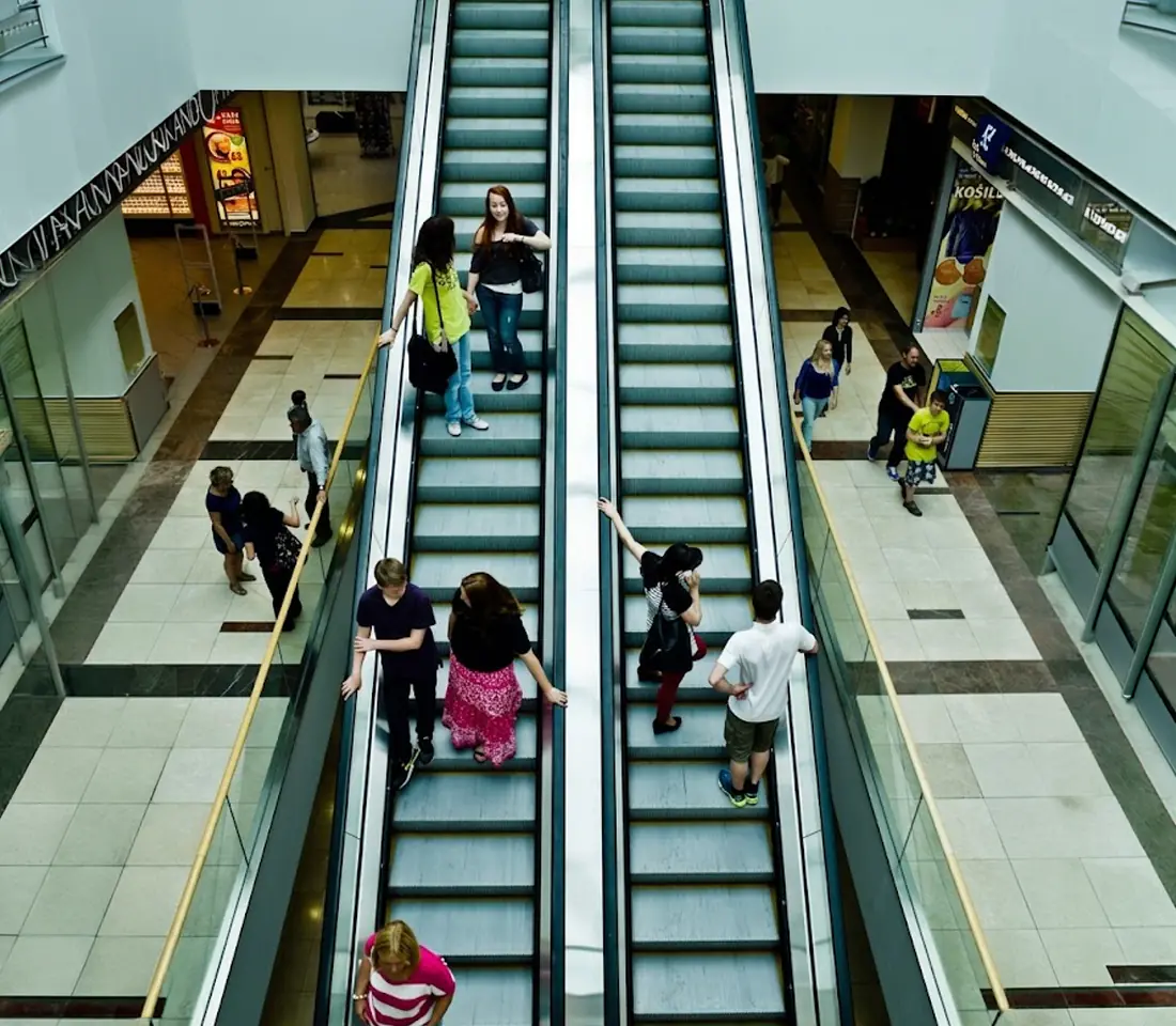 Low angle upward view of an escalator with yellow edged steps and glass side panels, receding toward a skylit ceiling.